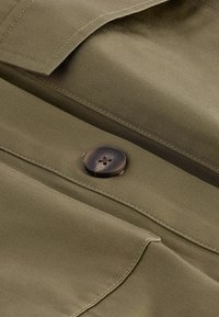 Nærbillede af khaki-stof med detaljeret syning og en hornlignende brun knap syet på en klap.