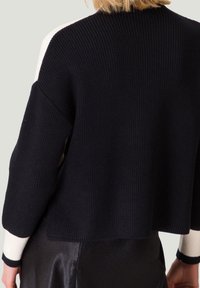 Pull en maille côtelée noir avec des accents blancs contrastants sur les épaules et les poignets, présentant une coupe décontractée et un tissu texturé.