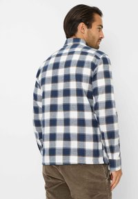 Camicia in flanella a quadri blu navy e bianca con dettagli marroni, dotata di collo e polsini abbottonati. Tessuto morbido, vestibilità rilassata.