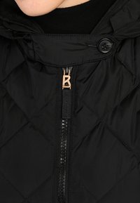 Veste matelassée noire avec capuche, dotée d'une fermeture éclair noire et d'un détail en bronze, mettant en valeur une finition texturée et lisse.