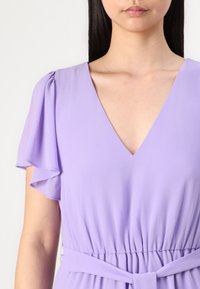 Lavanda haljina od lagane tkanine, s dubokim V-izrezom, lepršavim rukavima i vezanim pojasom za definiranu siluetu.