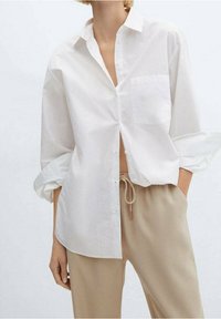 Camisa blanca de botones en algodón ligero, con un bolsillo en el pecho, corte relajado y mangas enrolladas, combinada con pantalones beige con cordón.