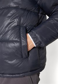 Donkerblauwe pufferjas met een glanzende afwerking, voorzien van een zijzak en elastische boorden. Het ontwerp bevat doorgestikte panelen en een losse pasvorm.