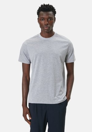 Joven con el pelo corto y rizado que lleva una camiseta de manga corta lisa de color gris claro y pantalones oscuros, de pie con una mano en el bolsillo.