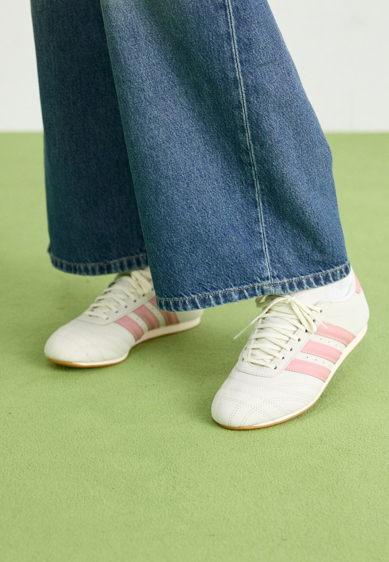 Bele športne copate z roza črtami, teksturirana usnja in ravnim gumijastim podplatom, v kombinaciji s širokimi jeans hlačami na zeleni tla.