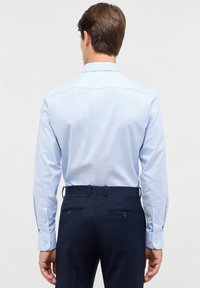 Man die met zijn rug naar de camera staat, gekleed in een lichtblauw overhemden met lange mouwen en donkerblauwe pantalons voor een effen achtergrond.