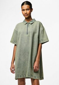 Traper haljina u prigušenoj zelenoj boji, s ovratnikom i kratkim rukavima. Opušteni kroj s zatvaračem, vidljivim šavovima i mekim teksturom.