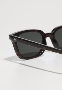 Gafas de sol carey con un marco angular audaz y lentes oscuros. Presenta un logo de Ray-Ban en plata en la patilla. Textura suave.
