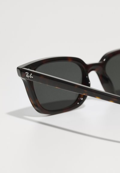 Lunettes de soleil écaille de tortue avec une monture angulaire audacieuse et des verres foncés. Présente un logo Ray-Ban argenté sur la branche. Texture lisse.