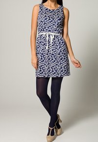 Abito senza maniche blu navy con motivo floreale bianco, stretta in vita con una cintura bianca, abbinato a collant scuri e scarpe beige.