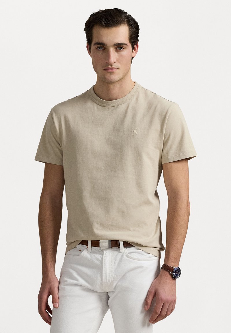 Beige bomull t-shirt med rund hals och korta ärmar, med en liten broderad logga på vänster bröst. Modellen bär vita byxor.