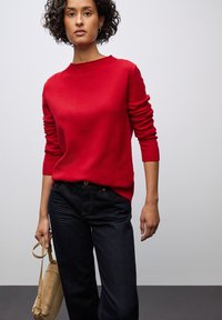 Maglione rosso a maglia con scollo rotondo e polsini a costine, abbinato a jeans in denim scuro. Il modello tiene una borsa in suede color sabbia.