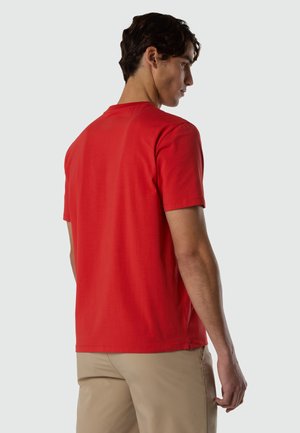 Mann, der nach links schaut, trägt ein einfaches rotes kurzärmliges T-Shirt und beige Hosen vor einem hellgrauen Hintergrund.
