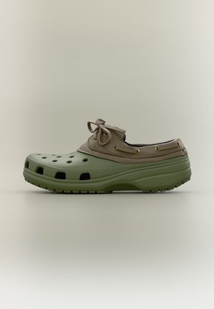 Sabot vert et beige unique avec des trous d'aération et un détail lacé, montré de côté sur un fond uni.