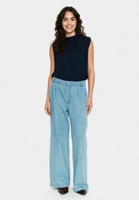 Top senza maniche blu navy con dettagli plissettati, abbinato a jeans a gamba larga blu chiaro. La modella sorride in piedi, indossando scarpe scure.