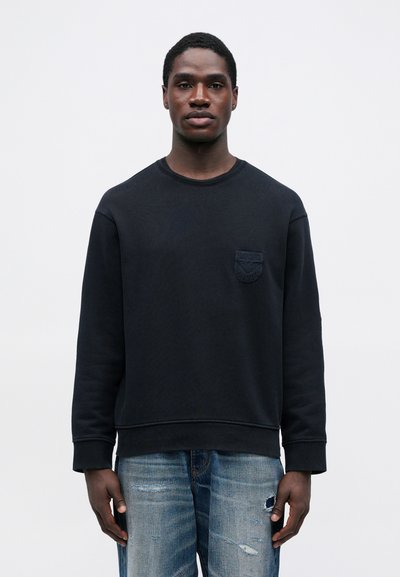 Νεαρός άνδρας φορώντας μια μαύρη μακρυμάνικη μπλούζα με λογότυπο στο στήθος και ελαφριά μπλε ξεθωριασμένα τζιν, στέκεται μπροστά σε έναν απλό λευκό τοίχο.