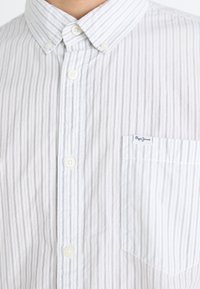 Camicia bianca con bottoni, caratterizzata da sottili righe verticali blu, colletto a punta, taschino frontale e etichetta del marchio sulla tasca del petto.