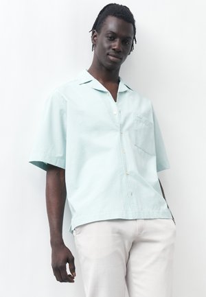 Νέος άνδρας με κοντά dreadlocks που φορά ανοιχτό μπλε πουκάμισο με κοντά μανίκια και λευκό παντελόνι, στέκεται μπροστά σε λευκό φόντο.