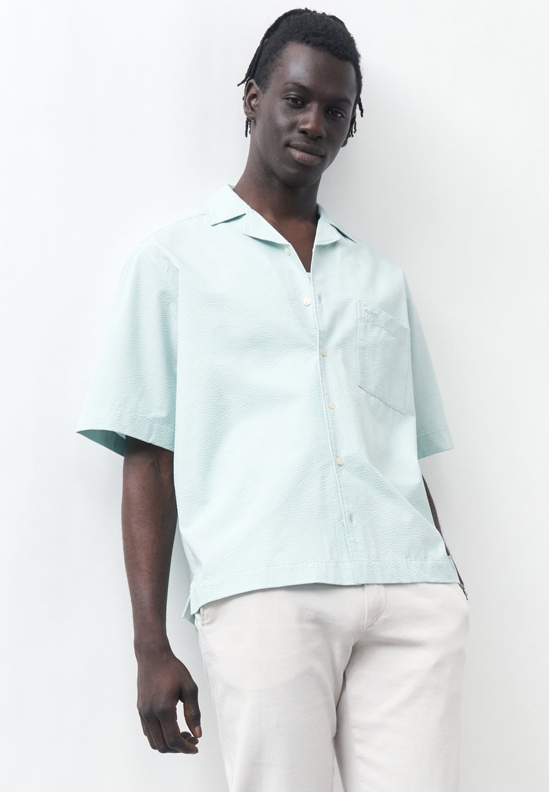 Νέος άνδρας με κοντά dreadlocks που φορά ανοιχτό μπλε πουκάμισο με κοντά μανίκια και λευκό παντελόνι, στέκεται μπροστά σε λευκό φόντο.