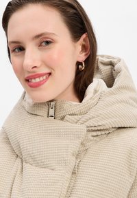 Manteau doudoune beige à capuche en tissu texturé. Doté d'une fermeture éclair à l'avant et d'un motif de quilting prononcé. Éléments de design minimalistes.