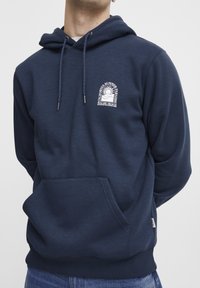 Marineblå hettegenser laget av mykt stoff, med en frontlomme, snorhette og et hvitt grafisk print på brystet.
