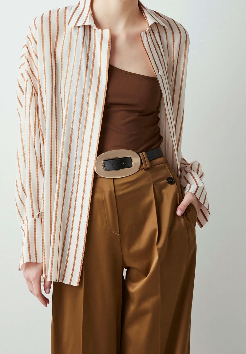 Lightweight, striped blouse in cream and rust, paired with a brown tank top and high-waisted, tan satin trousers featuring pleats and a belt.