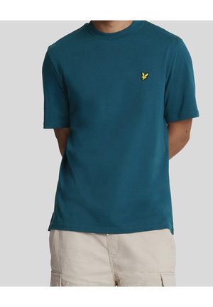 Mand iført en turkis t-shirt med korte ærmer og lille gult fuglelogo på brystet, kombineret med beige cargoshorts, står foran en grå baggrund.