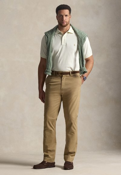 Homme debout portant un polo blanc, un pantalon kaki, une ceinture et des chaussures marron, avec un pull vert posé sur ses épaules.