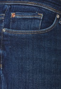 Tmavě modré denimové džíny s malou přední kapsou, měděnými a stříbrnými nýty, prošívanými detaily a hladkým povrchem.