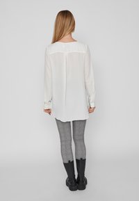 Blouse blanche à manches longues avec un col rond, en tissu doux, et une fente au dos, associée à un pantalon gris à carreaux et des bottes noires.