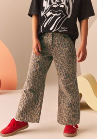 Pantaloni a stampa leopardata in beige e marrone, realizzati in denim, con una vestibilità a gamba larga. Abbinati a sneakers rosse con lacci.
