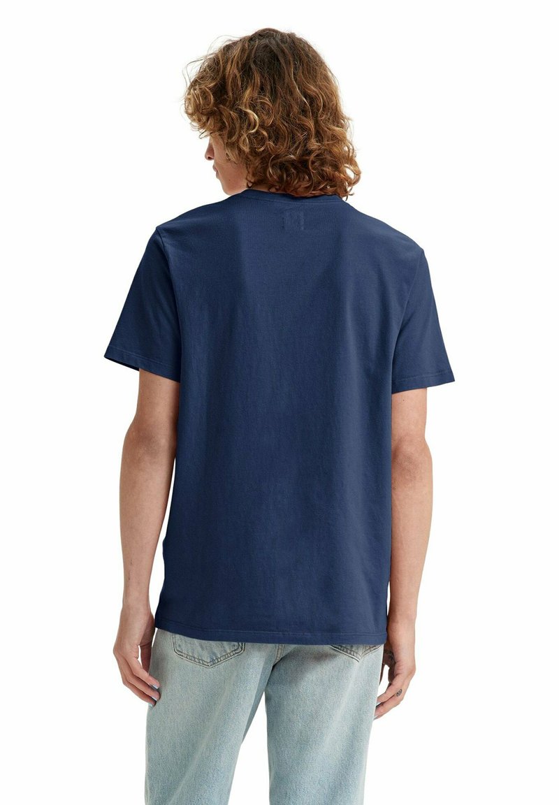 Person mit lockigem Haar, die ein einfarbiges dunkelblaues kurzärmliges T-Shirt und hellblaue Jeans trägt, mit dem Rücken zu einem weißen Hintergrund.