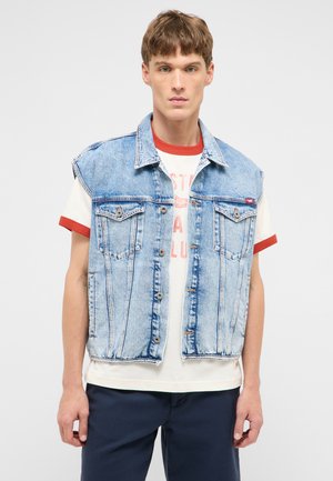Junger Mann, der eine hellblaue ärmellose Jeansweste über einem weißen T-Shirt mit roten Rändern und dunkler Hose trägt, steht vor einem einfarbigen Hintergrund.