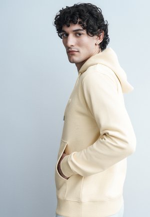 Ung mand med krøllet mørkt hår iført en beige hættetrøje, stående sidelæns med hænderne i lommerne mod en ensfarvet lysegrå baggrund.