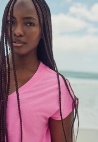 Ružičasta majica s V-izrezom od lagane tkanine, s kratkim rukavima i udobnim krojem. U pozadini je zamagljeni prizor obale.