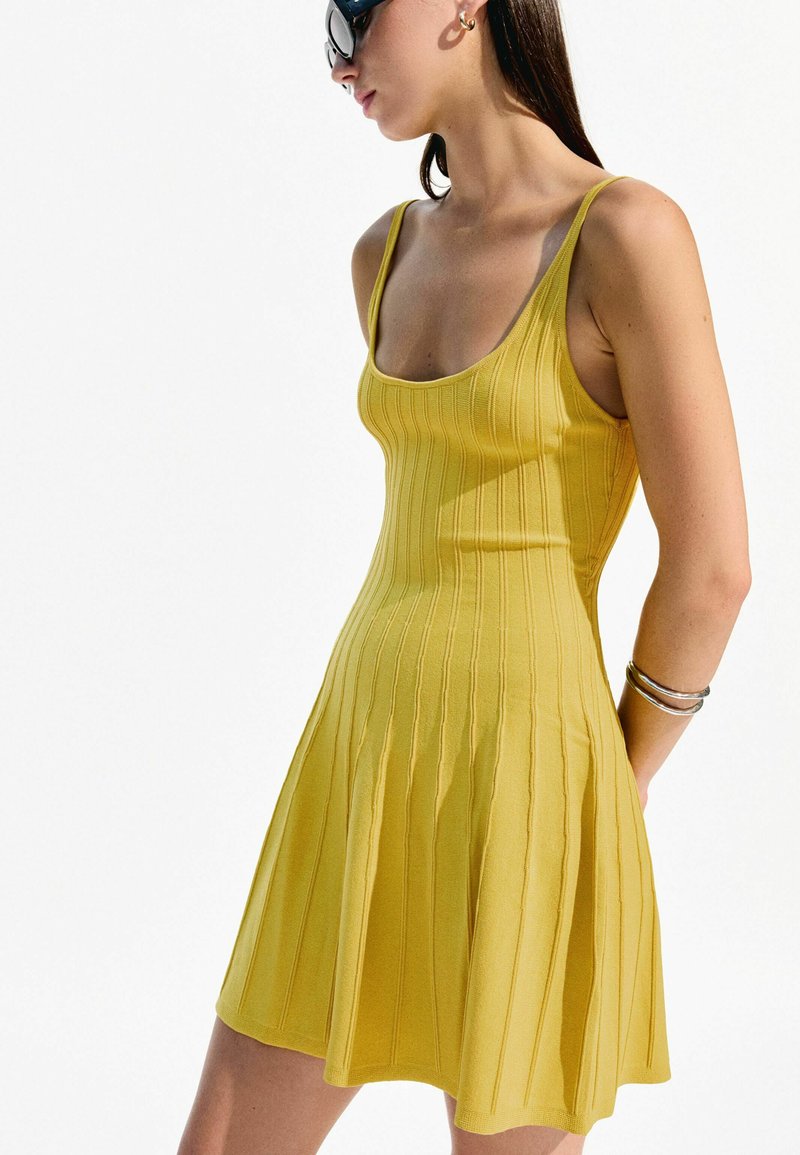 Abito giallo a coste con corpino aderente e gonna svasata, dotato di spalline sottili e delicate righe verticali, abbinato a bracciali d'argento.