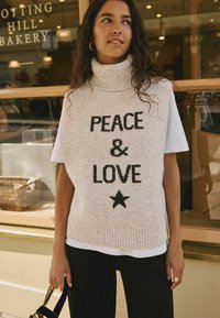Maglione senza maniche a collo alto in maglia beige con testo nero che recita "PEACE & LOVE" e motivo a stella, abbinato a una camicia bianca sotto.