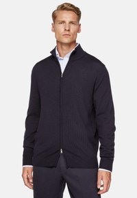 Navy zip-up sweater with a textured knit, ribbed cuffs, and collar. Features a slim fit and is paired with a light striped shirt underneath.