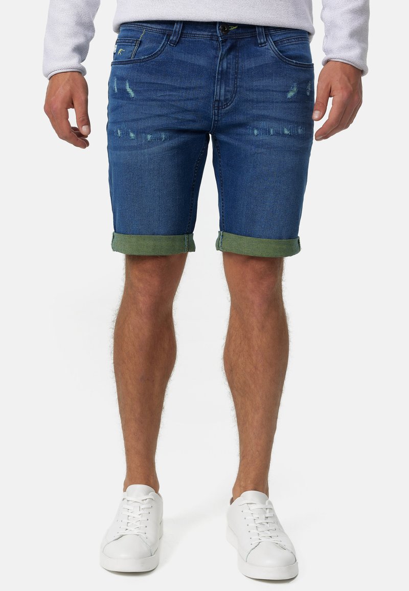 Donkere denim shorts met een vervaagd ontwerp en beschadigde details, met opgerolde groene boorden, gecombineerd met witte sneakers.