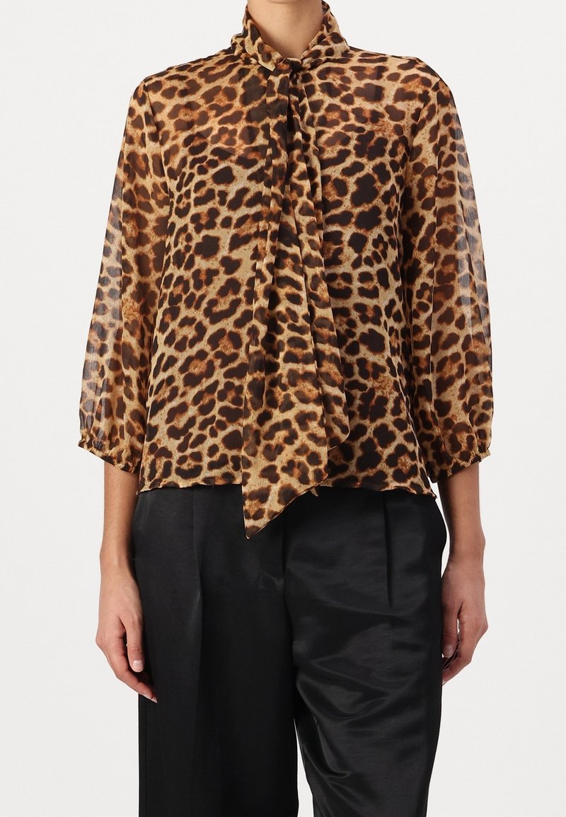 Blouse imprimée léopard en tissu léger et transparent avec un nœud au cou. Présente des manches trois-quarts et une coupe décontractée. Portée avec un pantalon noir.