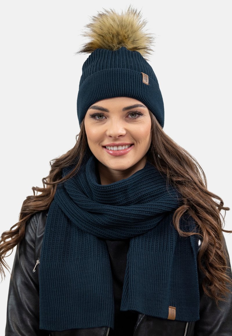 Dunkelblaues Strickmütze mit einem Fellpompon, kombiniert mit einem passenden strukturierten Schal. Verfügt über gerippte Muster und ein kleines Lederetikett als Akzent.