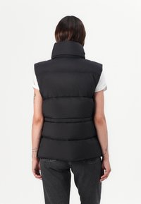 Svart boblevest med høy krage, ermeløs design, synlig quilting og glatt overflate. Brukt over en hvit skjorte sammen med mørkeblå jeans.
