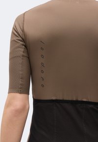 Κοντινό πλάνο στο άνω μέρος της πλάτης ενός ατόμου με καφέ και μαύρο ποδηλατικό μπλουζάκι με κοντό μανίκι και κατακόρυφο λογότυπο στο πλάι.