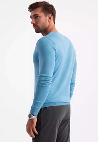 Maglione azzurro chiaro con maniche lunghe, polsini e orlo a coste. Indossato con pantaloni grigi scuri. Il tessuto appare morbido e liscio.