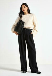 Donna che indossa un maglioneavoro a trecce, pantaloni neri a gamba larga, cintura nera, mocassini neri e porta una grande borsa tote in pelle nera.