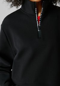 Oseba, oblečena v črno pulover s četrtinskimi zadrgami, na zadrgi piše "RED BULL RACING", delno viden rdeč kos oblačila pod njim.