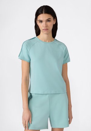 Jeune femme portant un haut à manches courtes bleu sarcelle clair et un short assorti, debout devant un fond blanc uni.