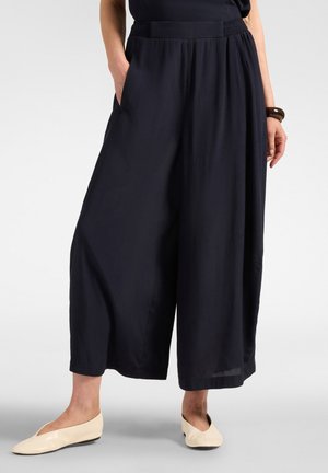 Personne portant un pantalon noir ample à jambes larges, mains dans les poches et chaussures plates couleur crème, debout sur un fond clair uni.