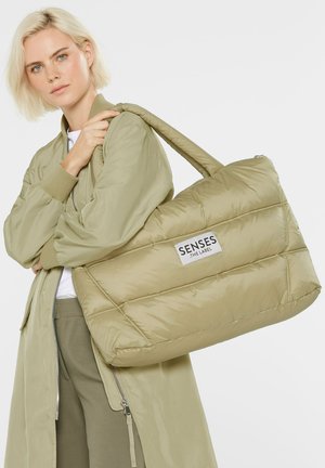 Padded tote bag in light olive green with a logo patch. Features thick straps and a quilted texture, complementing a bomber jacket.