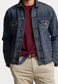 Muž ve modré džínové bundě přes vínový svetr, béžových kalhotách a hnědém opasku s modrožlutým pruhovaným koncem.